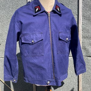 以下が含まれることがあります： ネイビーブルーのジャケット。フルレングスのジッパー、2つのフロントポケット、シルバーカラーのボタンが付いています。襟には赤と白のアクセントが付いた装飾パッチが付いています。マネキンに着せられています。