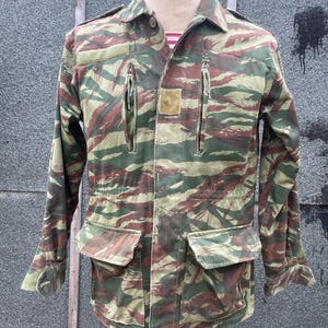 May include: A camouflage jacket in green, brown, and beige, with a tiger stripe design. The jacket has two chest pockets, two lower pockets, and two zipped pockets. It is buttoned up and displayed on a mannequin.