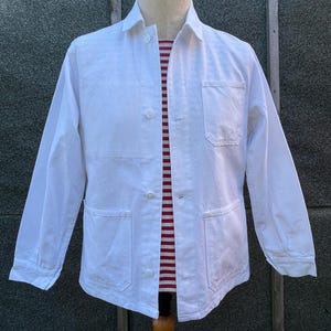 May include: A white work jacket with long sleeves, a collared neckline, and three front pockets. The jacket is buttoned up, revealing a red and white striped shirt underneath. The jacket is displayed on a mannequin.