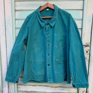 Puede incluir: Una chaqueta de trabajo azul turquesa con cierre de botones y varios bolsillos. La chaqueta está colgada de una percha de madera contra una puerta de madera verde claro.