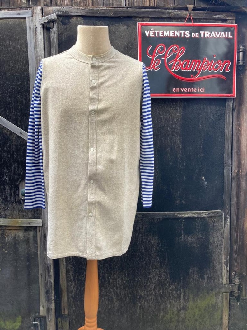 Puede incluir: Una camisa beige de manga larga con mangas a rayas azules y blancas. La camisa tiene un cuello mandar&iacute;n y un cierre de botones en la parte delantera. La camisa se muestra en un maniqu&iacute; sobre un fondo de madera.