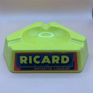 May include: A bright green ashtray with a hexagonal design. The ashtray displays the brand name "RICARD" in white letters on a blue background, with "ANISETTE LIQUEUR" underneath. It has a curved interior and a groove for cigarettes.
