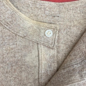 Puede incluir: Primer plano del cuello de una camisa beige con dos botones. La camisa est&aacute; hecha de una tela suave y texturizada.