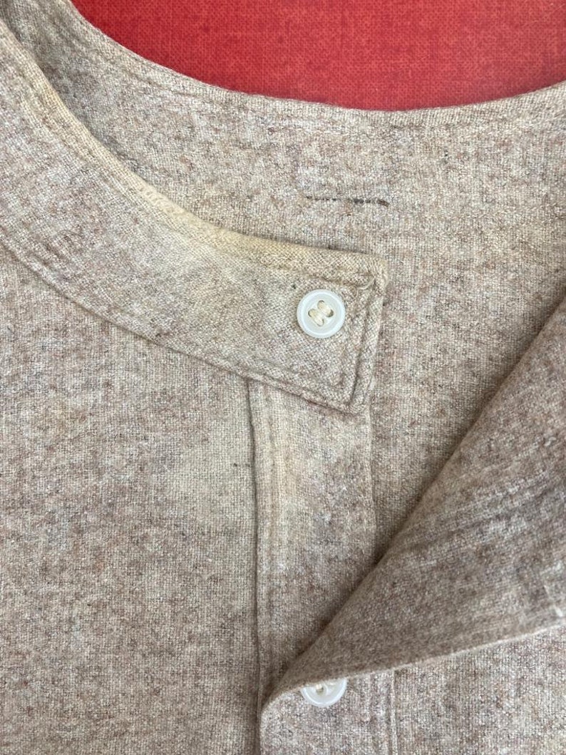Puede incluir: Primer plano del cuello de una camisa beige con dos botones. La camisa est&aacute; hecha de una tela suave y texturizada.