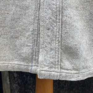 Puede incluir: Primer plano del borde inferior de una camisa de lino marr&oacute;n claro. La camisa tiene una sola costura en el centro delantero y una doble costura a lo largo del borde inferior.