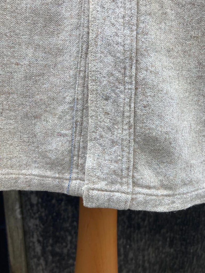 Puede incluir: Primer plano del borde inferior de una camisa de lino marr&oacute;n claro. La camisa tiene una sola costura en el centro delantero y una doble costura a lo largo del borde inferior.