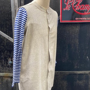 Puede incluir: Una camisa de lino beige con cuello mandar&iacute;n y cierre de botones en la parte delantera. La camisa tiene mangas largas y se lleva sobre una camisa a rayas azules y blancas.