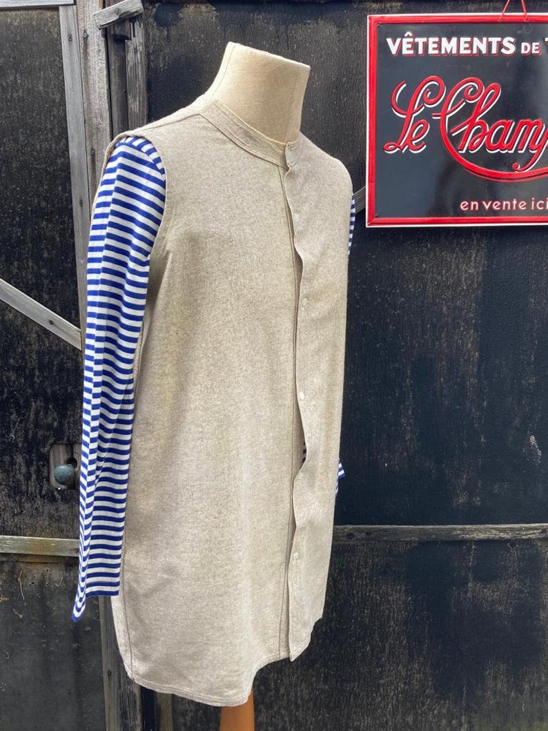 Puede incluir: Una camisa de lino beige con cuello mandar&iacute;n y cierre de botones en la parte delantera. La camisa tiene mangas largas y se lleva sobre una camisa a rayas azules y blancas.