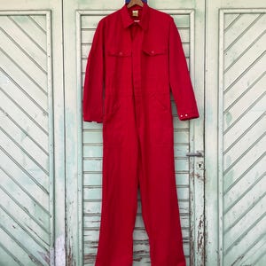 May include: A bright red long-sleeve jumpsuit featuring a collared neckline, a front button-down placket, two chest pockets with buttoned flaps, and long straight-leg trousers. The garment is displayed on a wooden hanger against a door.