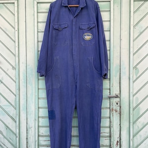 May include: A vintage blue work jumpsuit with long sleeves, multiple pockets, and a patch on the knee. The jumpsuit features a collared neckline and a small embroidered patch on the chest. The garment is hanging on a wooden hanger.