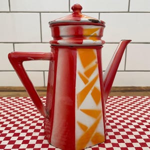 May include: A tall, square-shaped enamel coffee pot featuring a vibrant red and white color scheme with orange geometric accents. The pot includes a matching lid, a triangular handle, and a pointed spout, showing signs of wear on the edges.