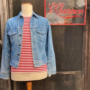 Op de afbeelding: Een blauwe denimjas met een rood-wit gestreept shirt eronder. De jas is op een mannequin voor een houten muur. Een vintage metalen bord met de tekst "VÊTEMENTS DE TRAVAIL Le Champion en vente ici" is op de muur.