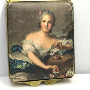 May include: A vintage gold-toned compact with a hinged lid. The compact features a colorful image of a woman in a white dress with a blue sash, holding flowers. The image is surrounded by a gold frame.