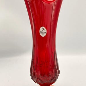 Puede incluir: Un jarrón de cristal rojo vibrante con un borde ondulado y una base acampanada. El jarrón tiene una forma delgada y alargada y un patrón texturizado cerca de la base. Una etiqueta blanca con texto negro es visible. El jarrón mide unos 25 cm de alto.