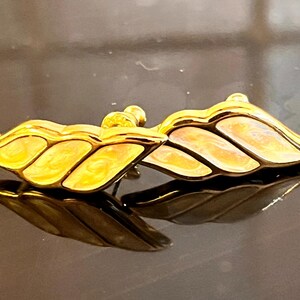 Puede incluir: Un par de pendientes dorados con forma de hoja. Los pendientes tienen un acabado brillante con un relleno de esmalte amarillo claro y melocotón. Los pendientes se muestran sobre una superficie reflectante.