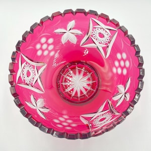 May include: A vibrant ruby-red glass bowl featuring detailed white starburst and floral patterns. The bowl has a scalloped edge and an intricate interior design. The design incorporates geometric shapes and floral motifs.