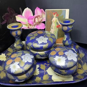 Puede incluir: Un juego de tocador azul y dorado con cuatro recipientes redondos y dos candelabros. Los recipientes tienen un diseño floral con flores blancas y detalles dorados. Los candelabros también están decorados con detalles dorados.