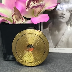 Puede incluir: Un espejo compacto dorado con un diseño decorativo y un pequeño centro redondo de color naranja. El espejo está sobre una superficie gris con estampado.