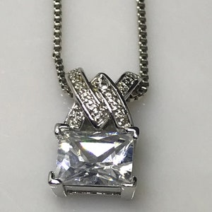 May include: A silver necklace with a square-shaped pendant featuring a large clear cubic zirconia stone. The pendant is accented with smaller cubic zirconia stones.