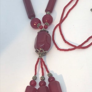 Puede incluir: Un collar largo de cuentas rojas con un gran colgante de piedra roja facetada. El colgante tiene un borla de plata con tres cuentas rojas más pequeñas y tres cuentas rectangulares rojas.