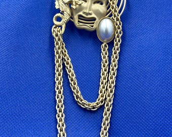May include: A gold-toned brooch featuring a theatrical mask with a pearl accent and dangling chains. The design includes a dragon-like figure and coiled details, set against a blue background.