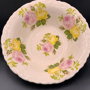 Può includere: Una ciotola rotonda in ceramica bianco sporco con bordo smerlato. La ciotola è decorata con rose gialle e rosa e foglie verdi. Le rose sono disposte attorno al bordo e al centro.