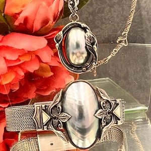 Puede incluir: Un collar de plata con un colgante y una pulsera a juego. El colgante y la pulsera presentan una piedra ovalada e iridiscente rodeada por un marco plateado decorativo con detalles de hojas. La pulsera tiene una banda de malla.