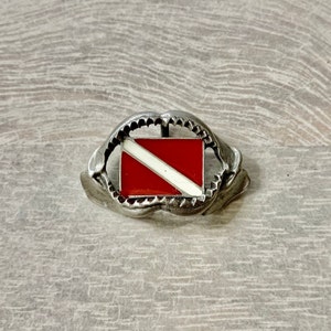 May include: A silver belt buckle with a red and white scuba diving flag in the center. The buckle is shaped like a shark's mouth.