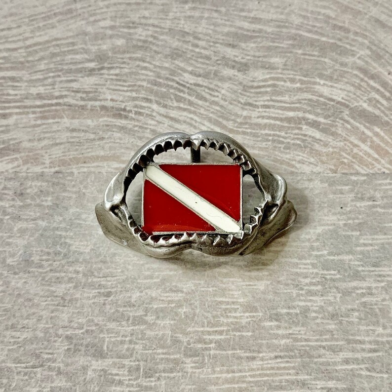 May include: A silver belt buckle with a red and white scuba diving flag in the center. The buckle is shaped like a shark's mouth.