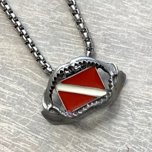 May include: A silver chain necklace with a silver pendant featuring a red and white scuba diving flag. The pendant is shaped like a shark's mouth.