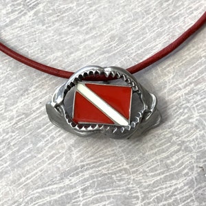 May include: A silver necklace with a red and white scuba diving flag pendant. The pendant is shaped like a shark's mouth.
