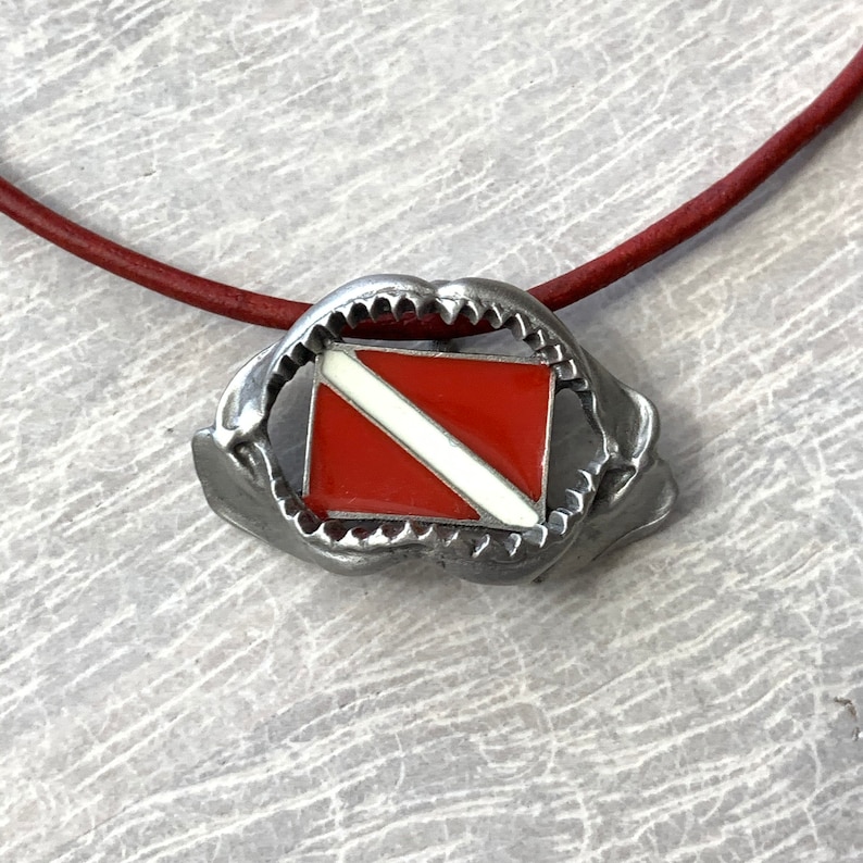 May include: A silver necklace with a red and white scuba diving flag pendant. The pendant is shaped like a shark's mouth.