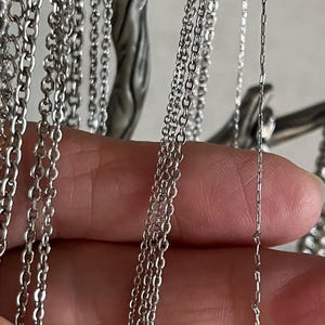 Pode incluir: Um close-up de várias correntes prateadas de diferentes espessuras, penduradas em um acessório de metal. As correntes estão dispostas em linha vertical, com a corrente mais fina à direita e a mais grossa à esquerda.