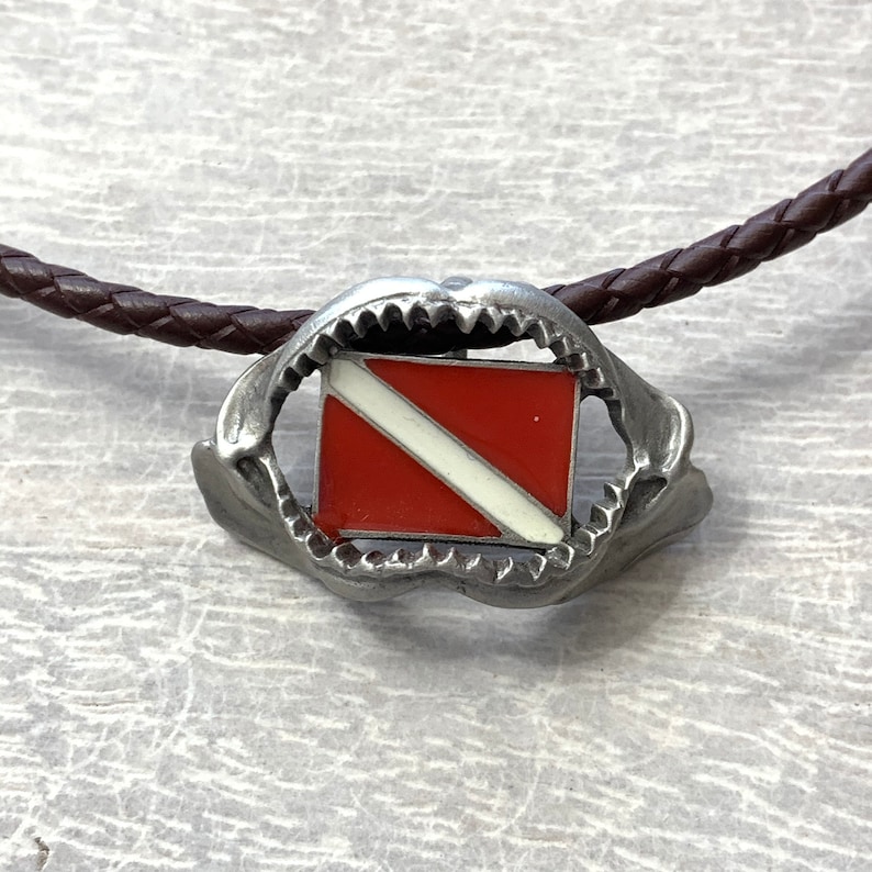 May include: A silver shark tooth pendant necklace with a red and white scuba diving flag inside the tooth. The necklace is on a brown leather cord.