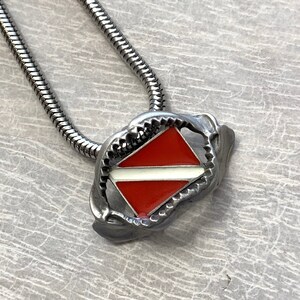 May include: A silver chain necklace with a silver pendant shaped like a shark's mouth holding a red and white scuba diving flag.