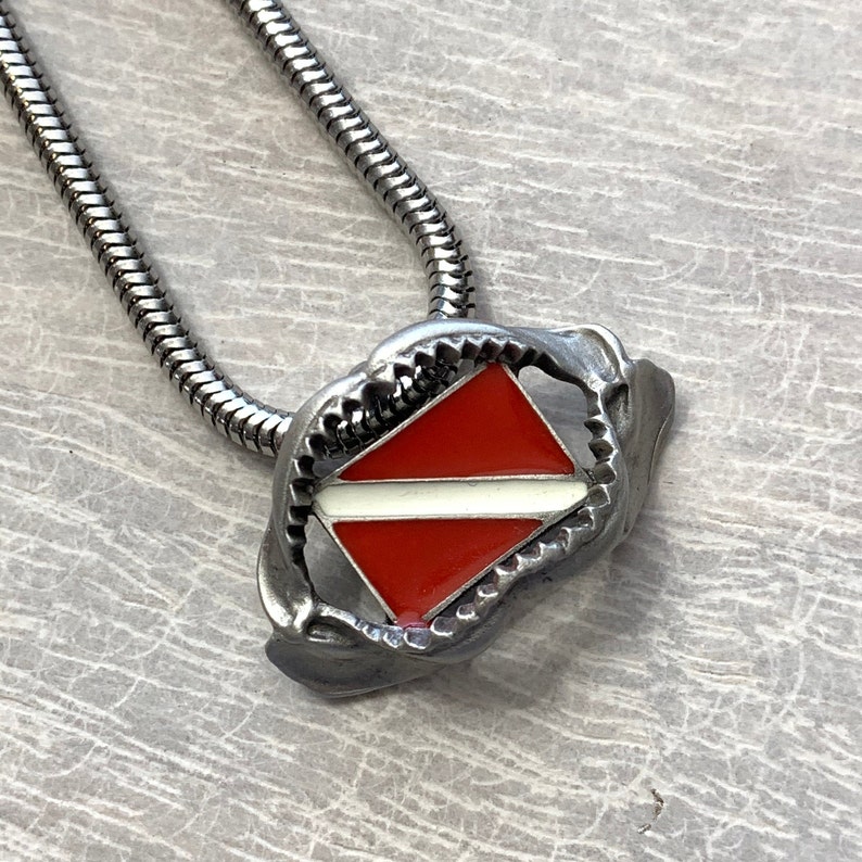 May include: A silver chain necklace with a silver pendant shaped like a shark's mouth holding a red and white scuba diving flag.