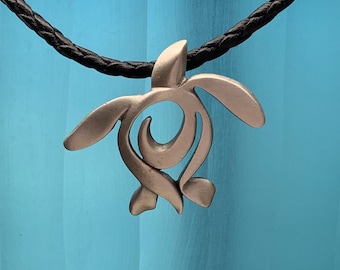 May include: A silver-toned turtle pendant necklace with a detailed, abstract design. The necklace is on a black cord.
