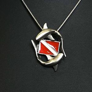 Puede incluir: Collar de plata con un colgante que representa dos tiburones plateados que rodean una bandera de buceo roja y blanca.