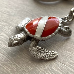 May include: A silver turtle keychain with a red and white enamel shell. The turtle is facing right and has detailed features.
