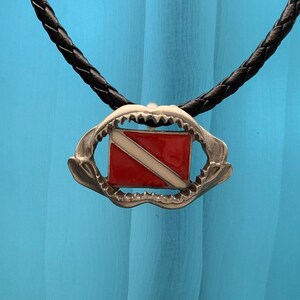 May include: A silver shark tooth pendant necklace with a red and white scuba diving flag inside. The necklace is on a black braided cord.