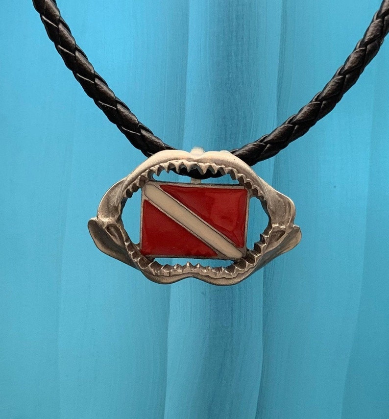 May include: A silver shark tooth pendant necklace with a red and white scuba diving flag inside. The necklace is on a black braided cord.