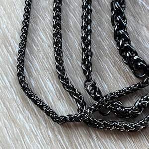 May include: Three black metal chains of different widths and styles. The chains are intertwined and laid out on a light wood surface.