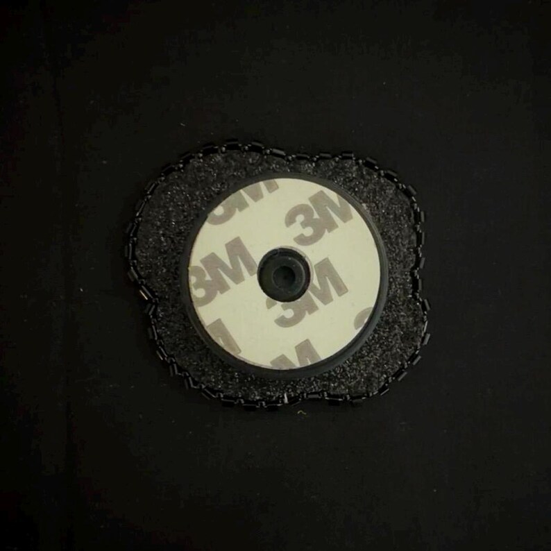 May include: A circular phone grip with a 3M adhesive backing. The grip is white with the 3M logo printed on it. It is surrounded by a black, textured border. The grip is designed to attach to the back of a phone.