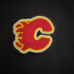 May include: A beaded patch featuring the Calgary Flames logo. The logo is a stylized red 'C' with a yellow outline and a black border, filled with red beads. The patch is surrounded by a black beaded border.