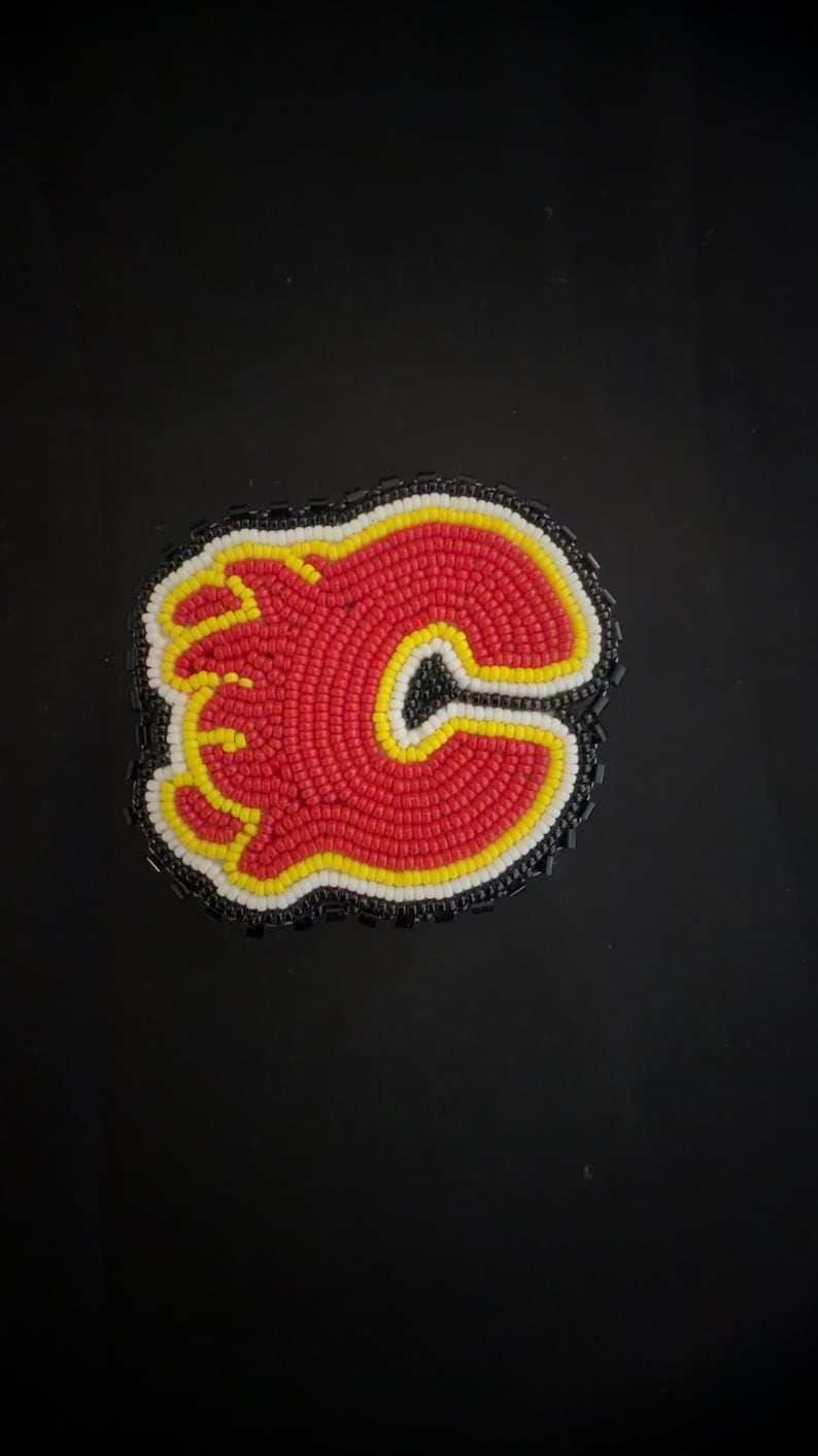 May include: A beaded patch featuring the Calgary Flames logo. The logo is a stylized red 'C' with a yellow outline and a black border, filled with red beads. The patch is surrounded by a black beaded border.
