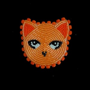 May include: Orange beaded cat face pin with black and light blue eyes. The cat pin is made of small, round beads and has a detailed design. The pin is set against a black background.