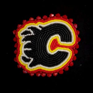 May include: A beaded patch featuring the Calgary Flames logo. The logo is a stylized "C" with a flame design, rendered in black, red, yellow, and white beads. The patch is outlined with red beads against a black background.