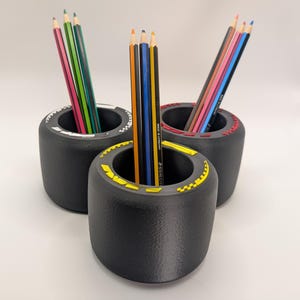 May include: Three black pencil holders shaped like racing tyres, each with a different coloured trim: white, yellow, and red. Pencils of various colours are inside each holder, creating a vibrant contrast. The holders are on a white surface.