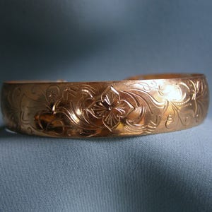 May include: A gold-colored bangle bracelet with an intricate floral pattern. The bracelet features a central flower design surrounded by swirling leaves and vines. The metal has a polished finish, reflecting light.