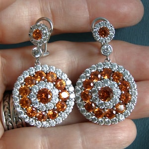 May include: A pair of silver-toned earrings with a circular design. Each earring features a central cluster of orange gemstones surrounded by a ring of white gemstones. The earrings have a post and clip closure.